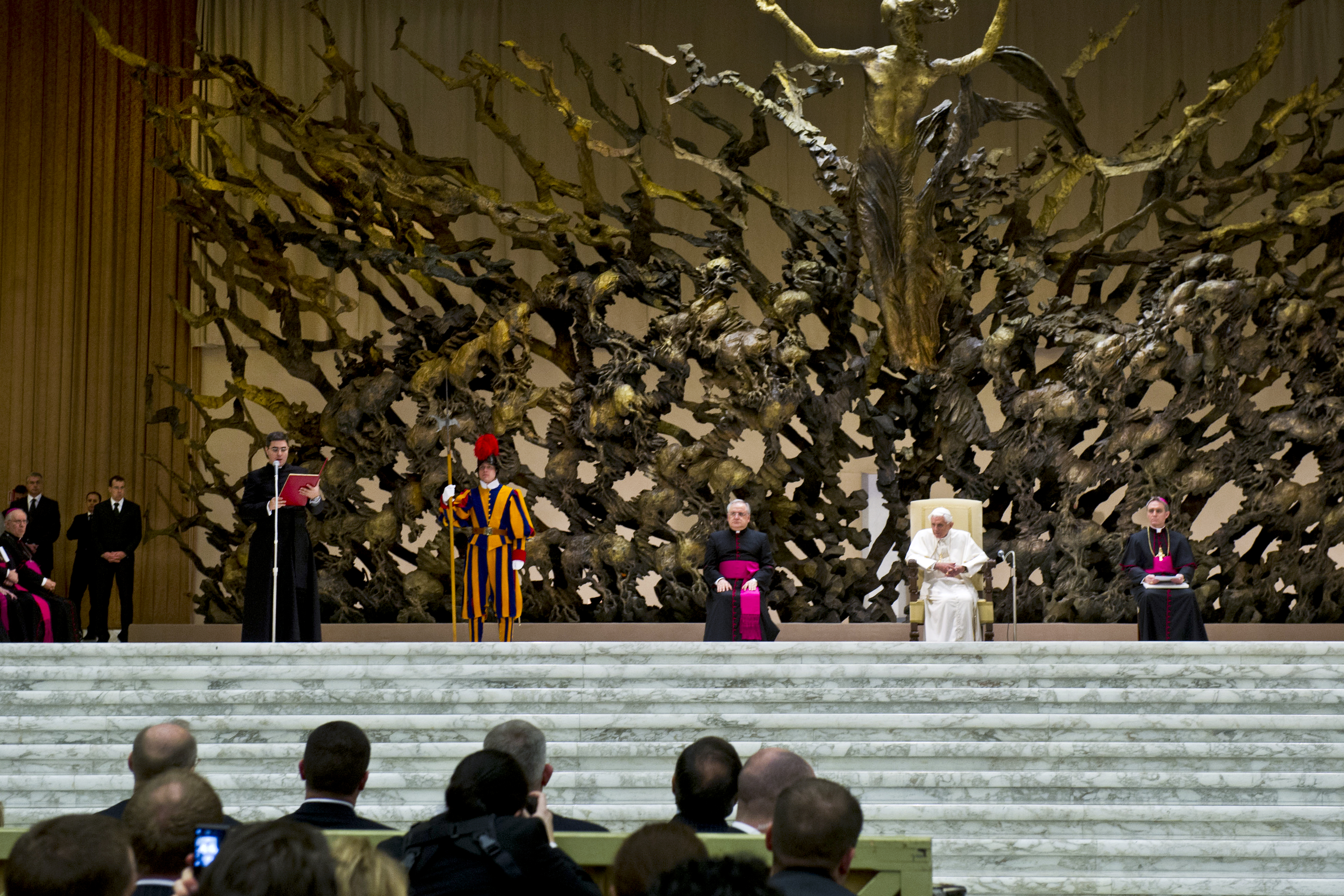 Het sculptuur domineert het podium van de Paulus VI-aula. Hier een foto uit januari 2013 met paus Benedictus XVI. © Wikimedia Het sculptuur domineert het podium van de Paulus VI-aula. Hier een foto uit januari 2013 met paus Benedictus XVI. © Wikimedia