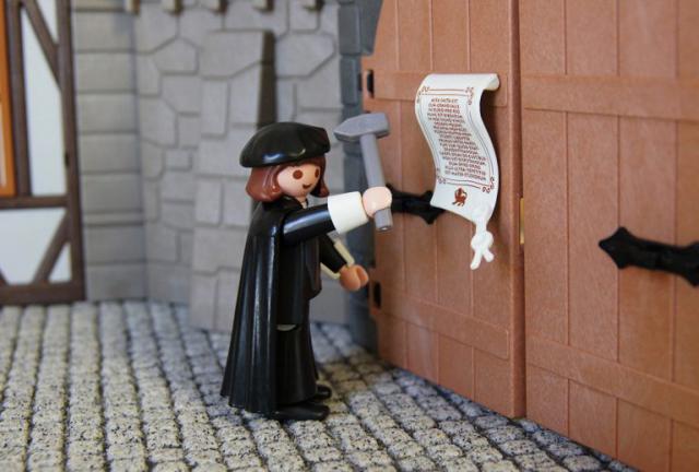 Een Lutherfiguur van Playmobil spijkert zijn 95 stellingen tegen de poort van de kerk in Wittenberg. 